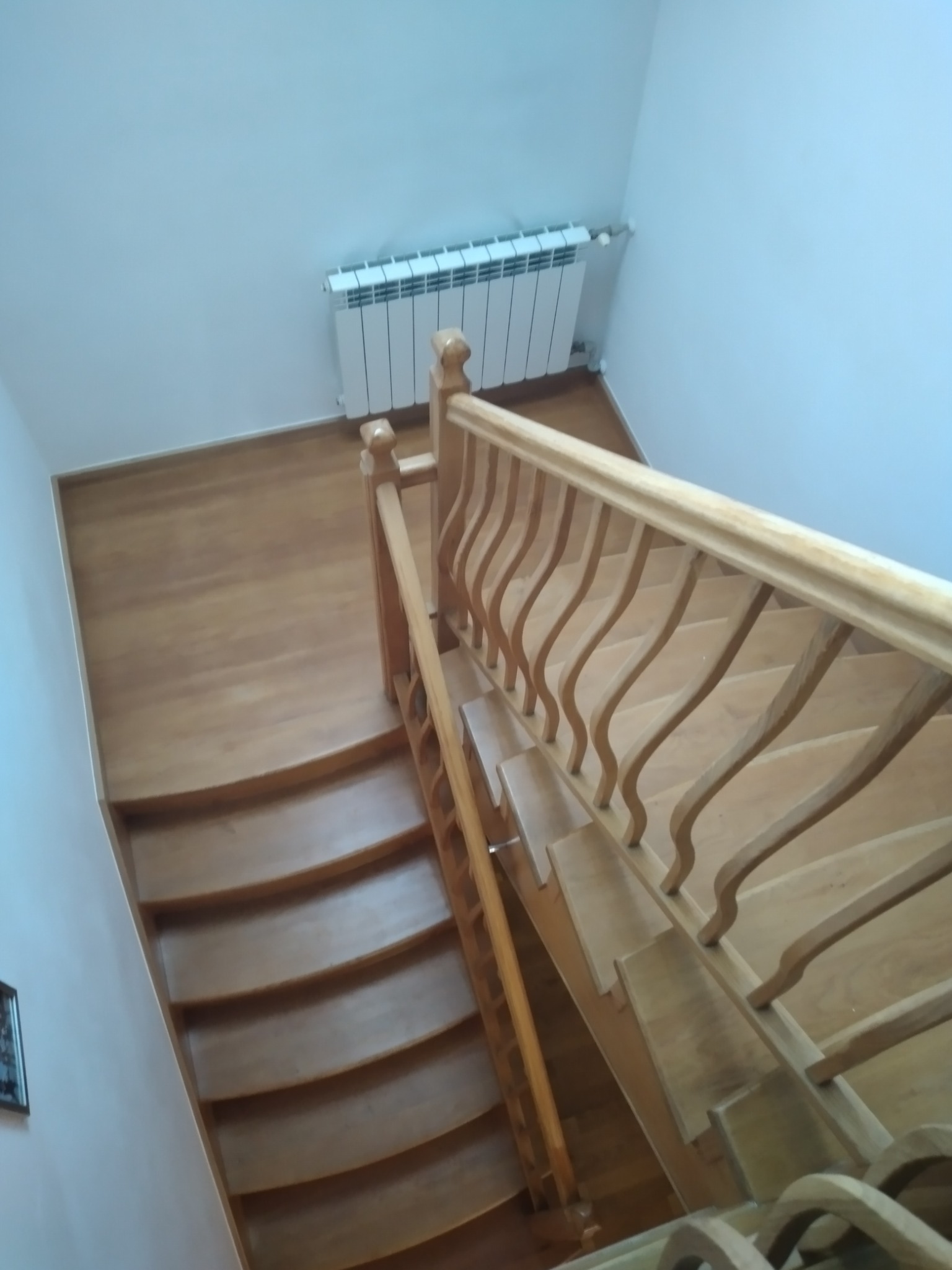 Widok z góry na drewniane schody z ozdobną balustradą w jasnym wnętrzu, z widocznym grzejnikiem na ścianie.