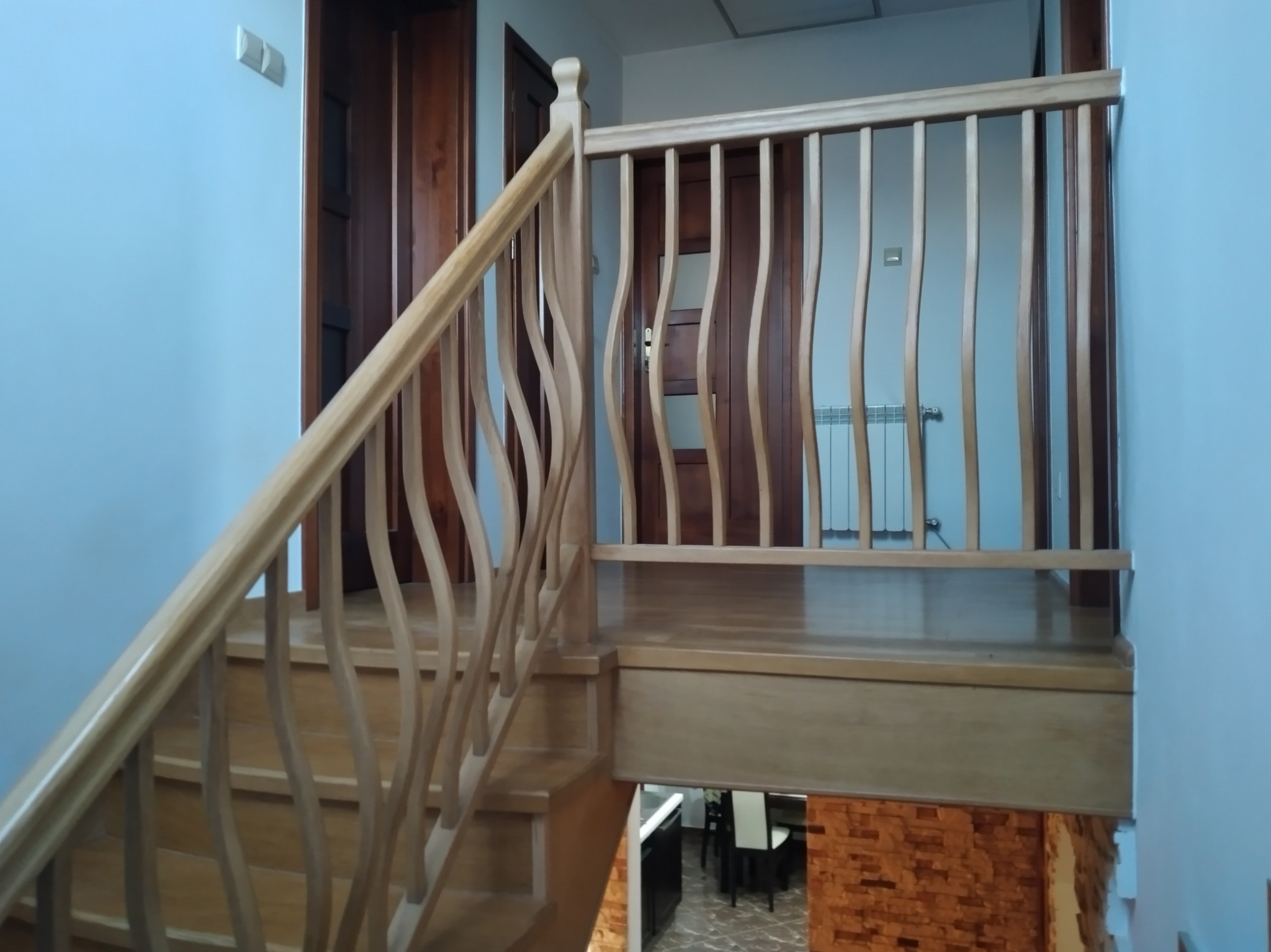 Jasne drewniane schody z falistą balustradą, widok z dołu, w tle drzwi i grzejnik.