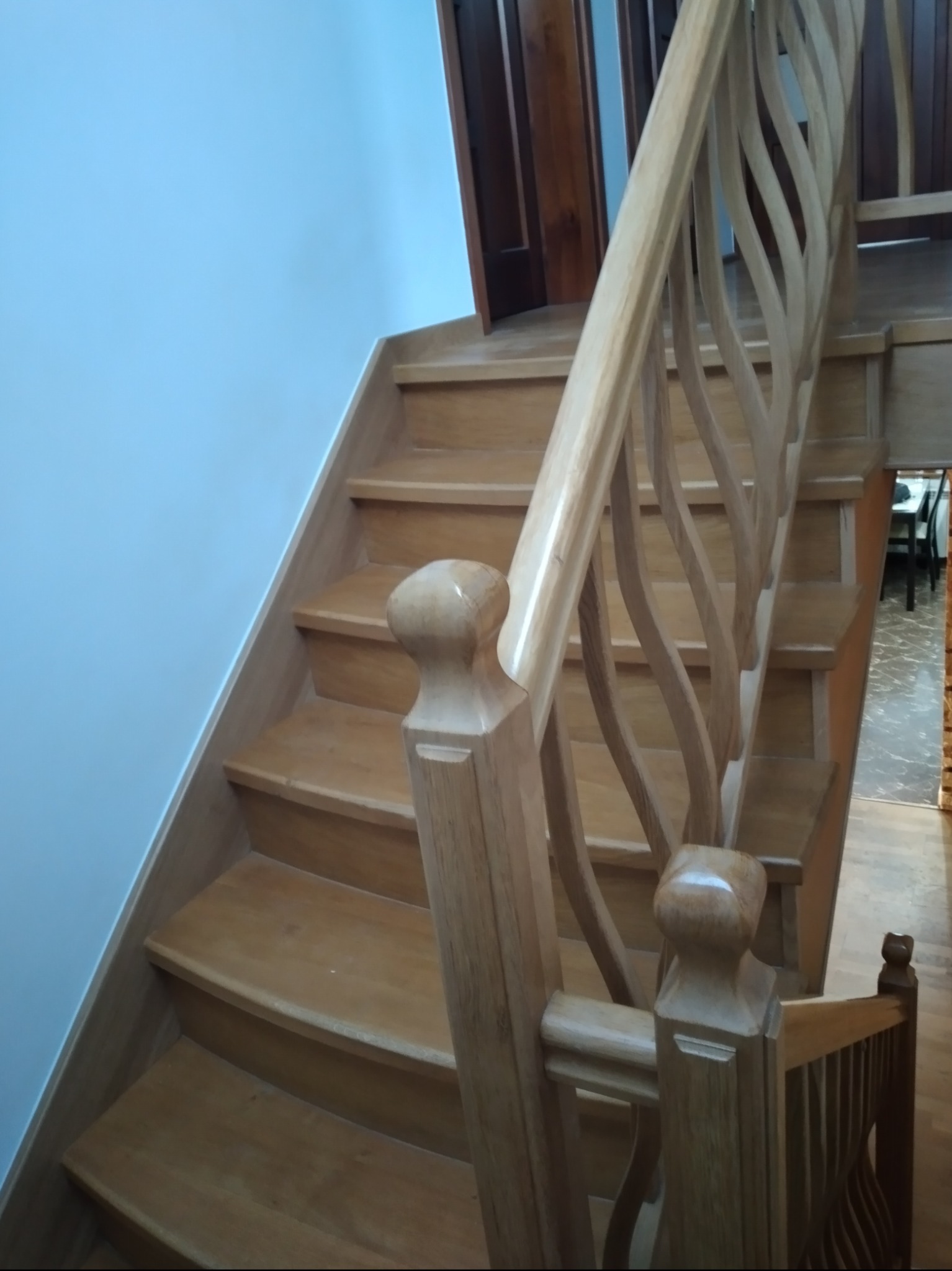 Jasne, drewniane schody z ozdobną, falistą balustradą, widok z dołu, niebieska ściana w tle.