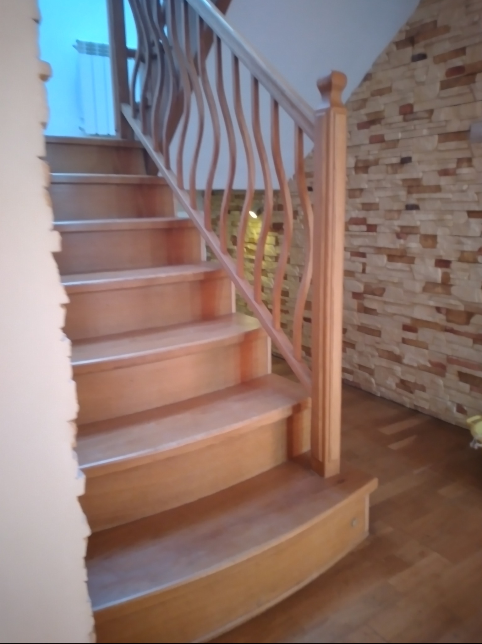 Jasne, drewniane schody z ozdobną, falowaną balustradą wewnątrz domu z kamienną ścianą w tle.