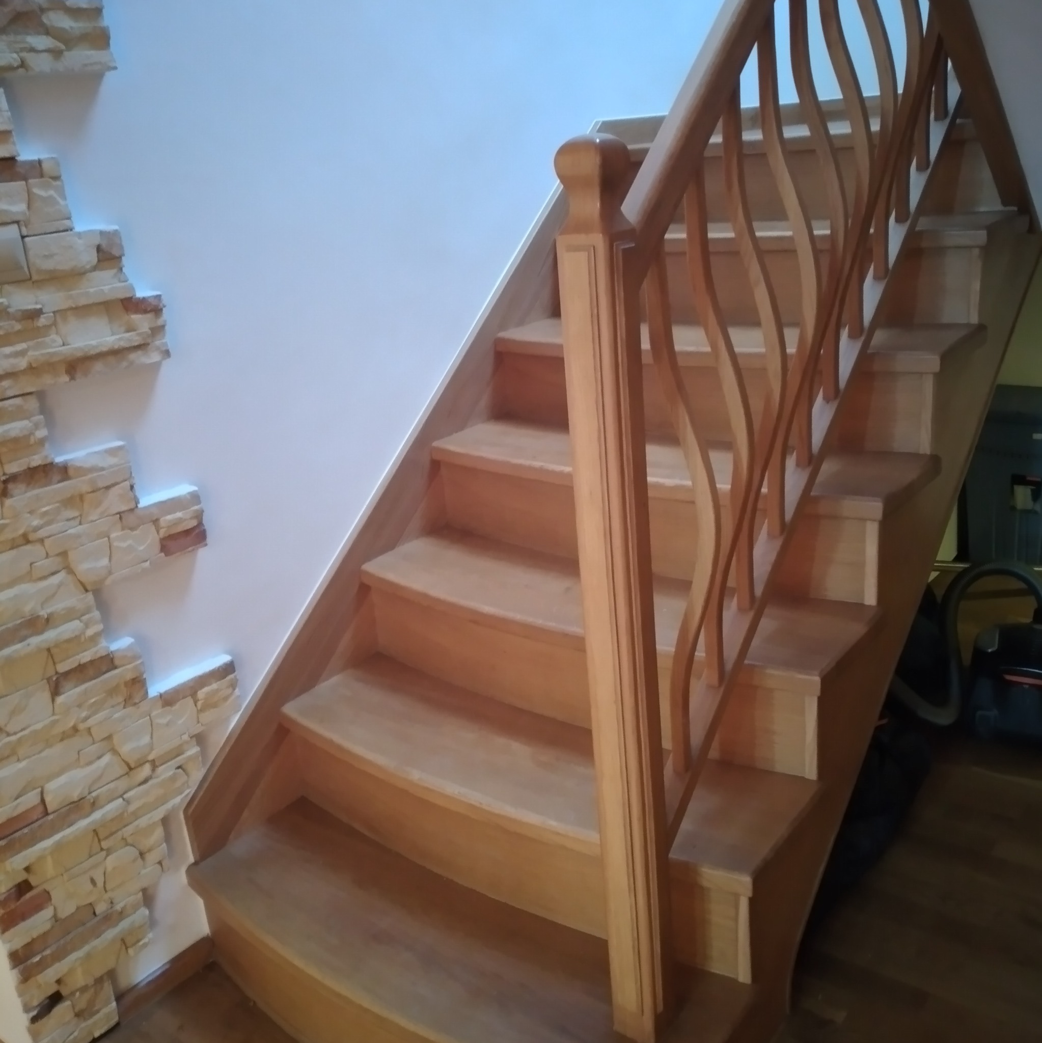 Drewniane schody z balustradą o falistym wzorze, wykończone jasnym drewnem, z dekoracyjną ścianą z kamienia w tle.