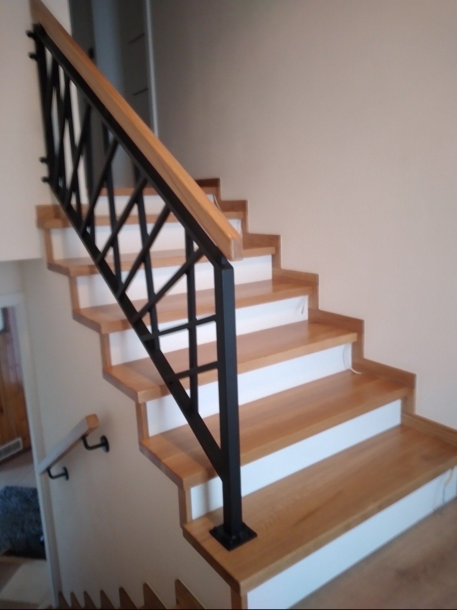 Jasne, drewniane stopnie schodów z białą podstopnicą i czarną, geometryczną balustradą. Dodatkowa poręcz na ścianie.