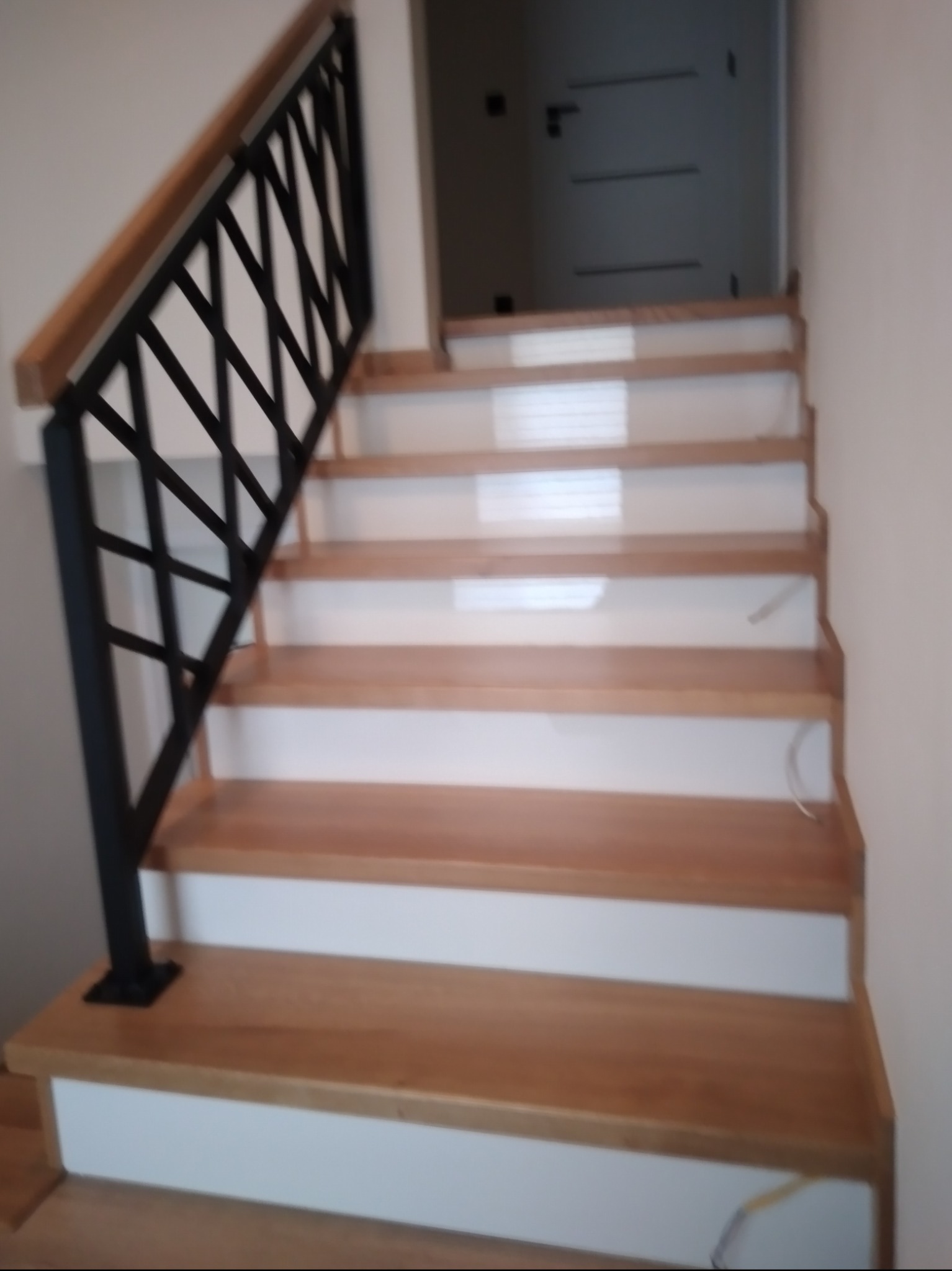 Wewnętrzne schody z drewnianymi stopniami i białą podstopnicą, z czarną, metalową balustradą o geometrycznym wzorze i drewnianym poręczą.