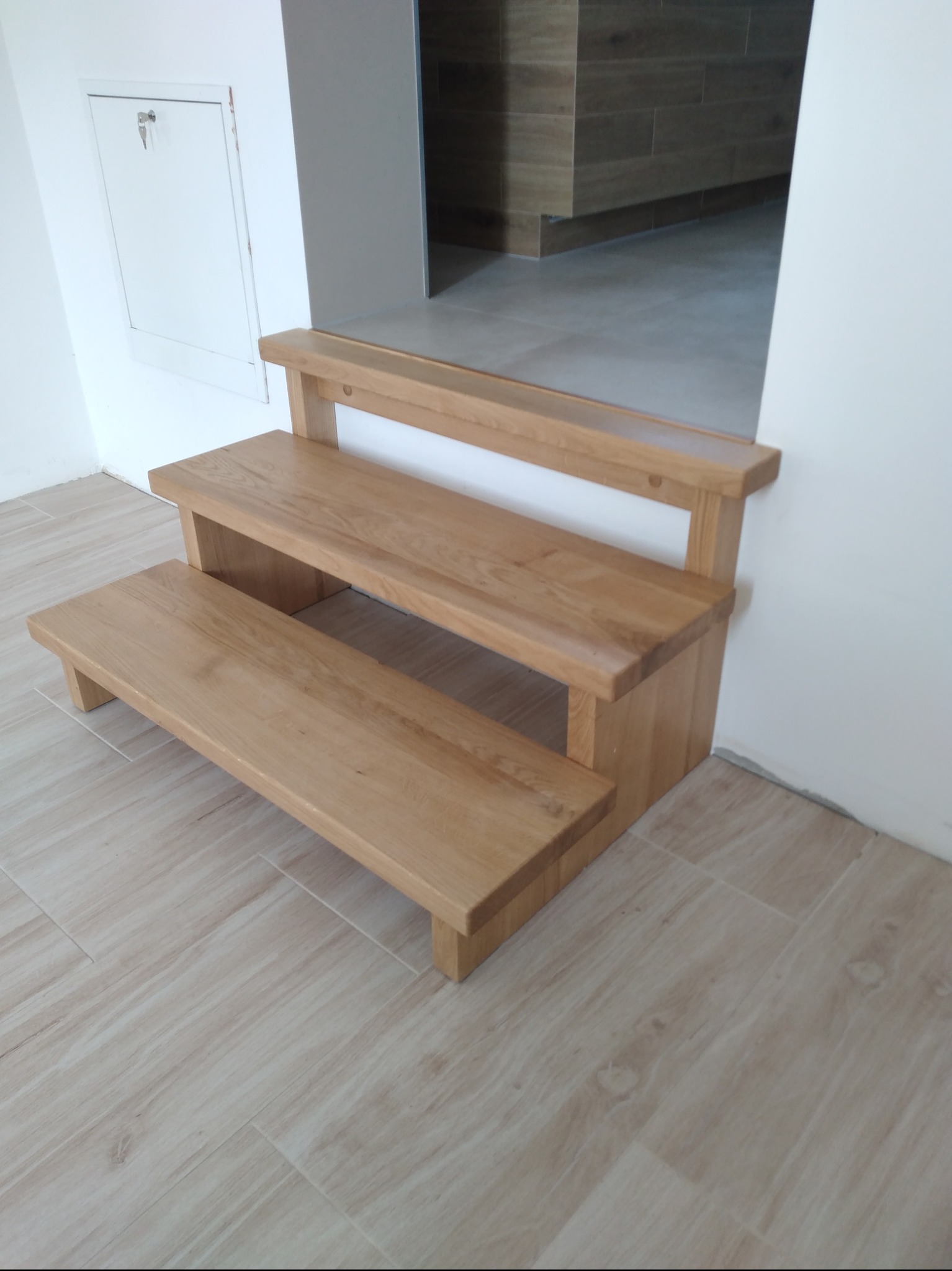 Krótkie, dwustopniowe schody z jasnego drewna, prowadzące do wyższego poziomu podłogi wyłożonej płytkami imitującymi drewno, z widoczną białą szafką z kluczem na ścianie po lewej stronie.