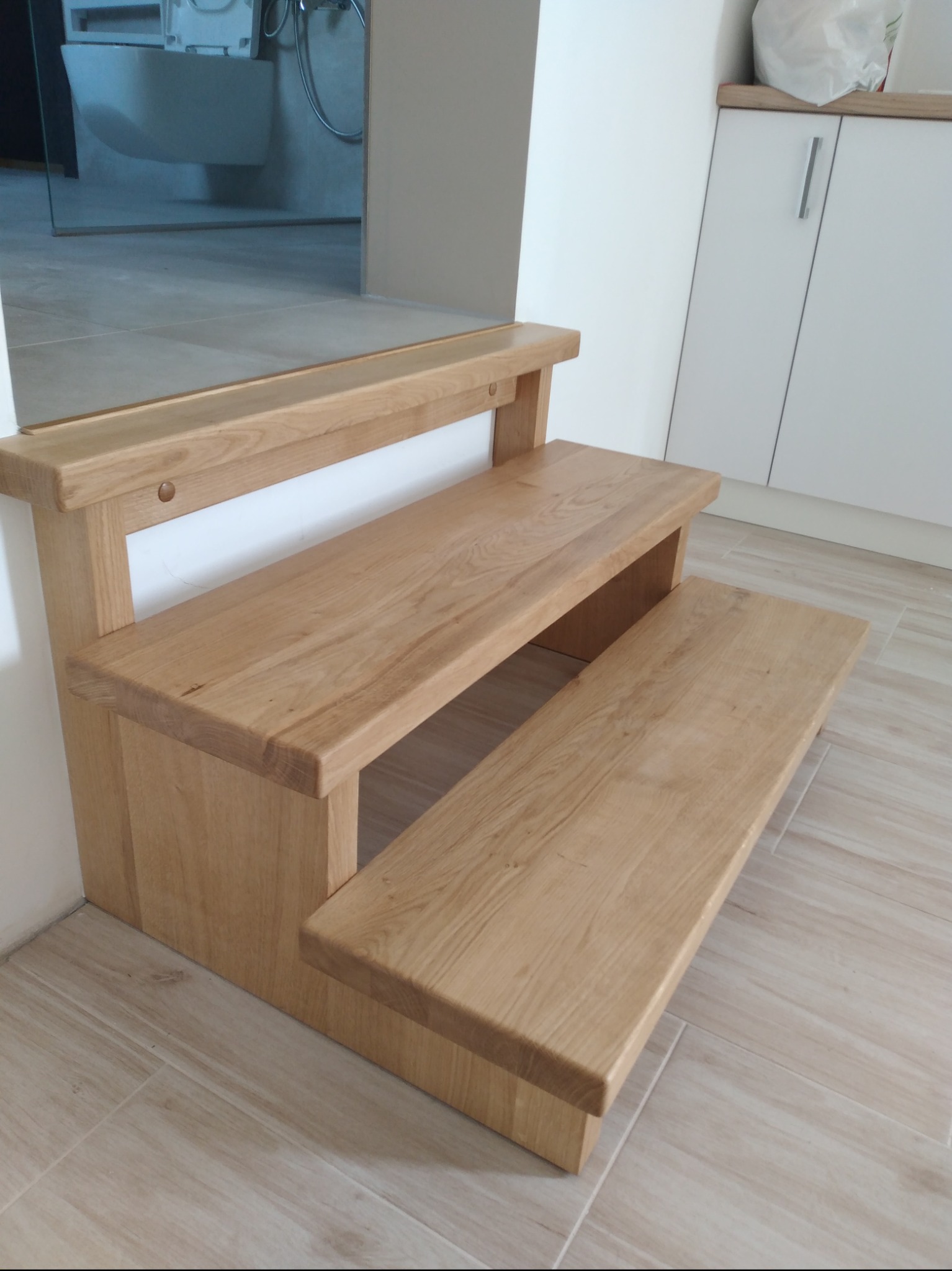 Niewielkie, dwustopniowe schodki drewniane z jasnego drewna dębowego, prowadzące do podwyższonego poziomu w łazience, z widoczną toaletą w tle.