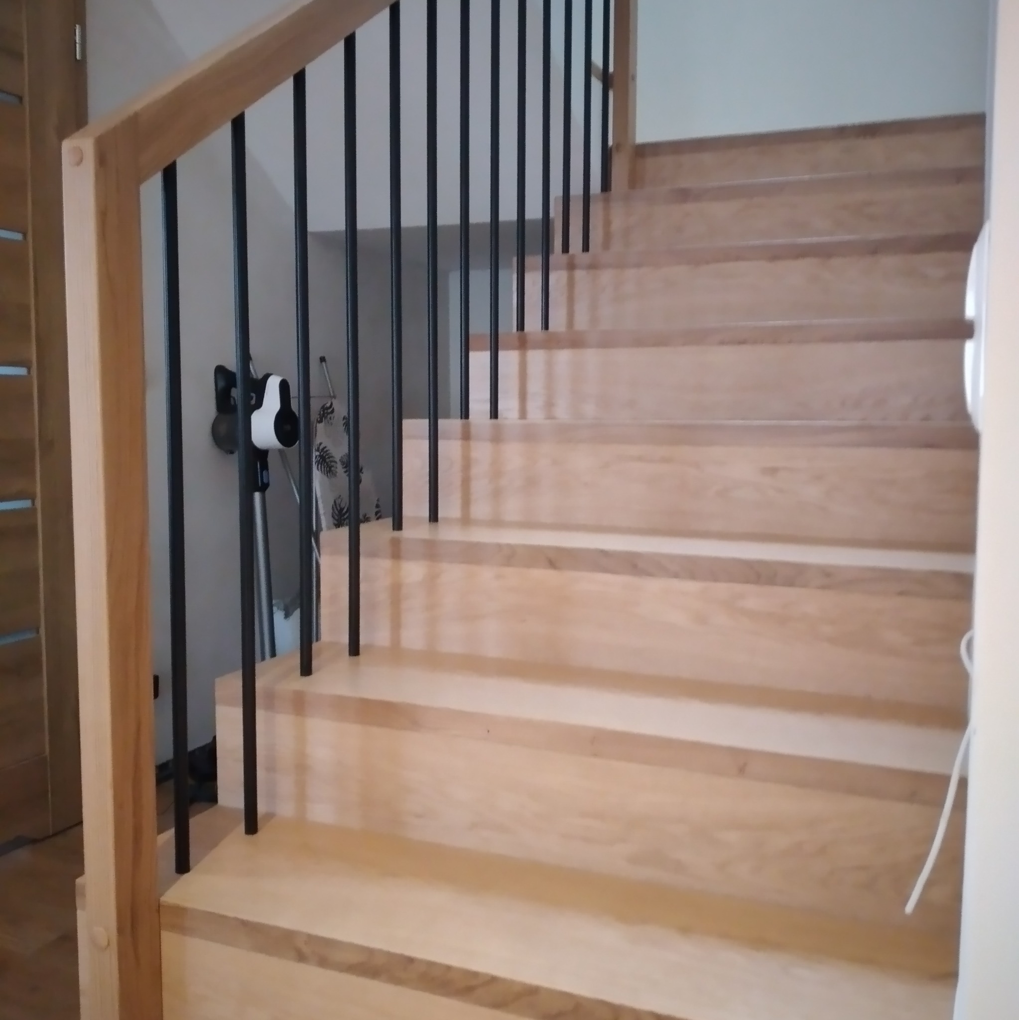 Jasne drewniane schody z czarnymi metalowymi balustradami w nowoczesnym wnętrzu, widoczny odkurzacz pionowy w tle.