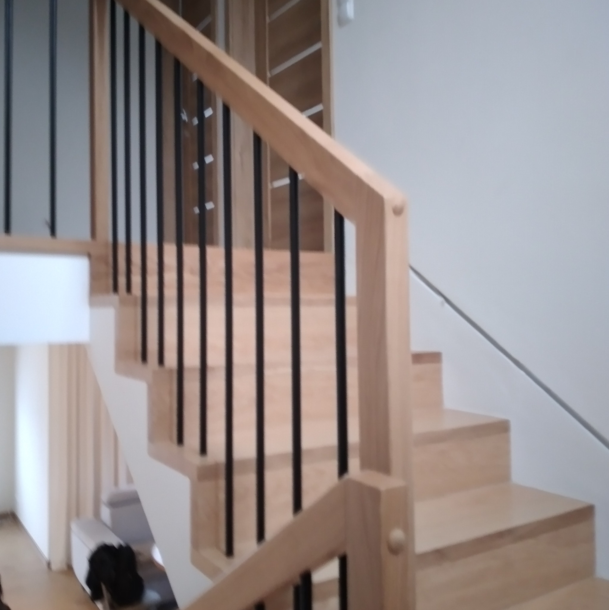 Jasne drewniane schody z czarnymi, pionowymi balustradami, widok z dołu, z fragmentem białej ściany i mebli w tle.