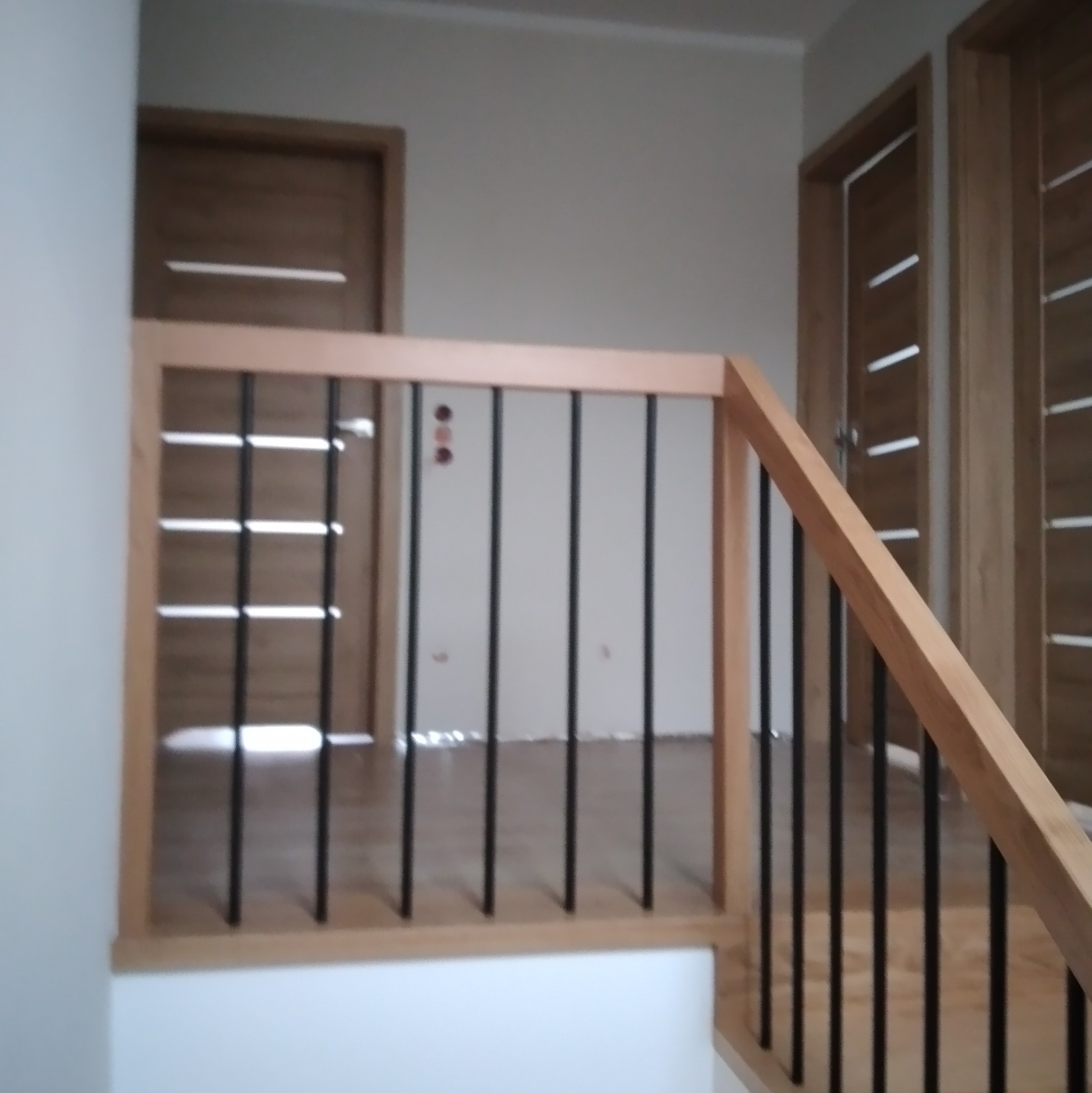 Fragment klatki schodowej z drewnianą balustradą o jasnym odcieniu i czarnymi, pionowymi szczebelkami, widoczne drzwi w tle.