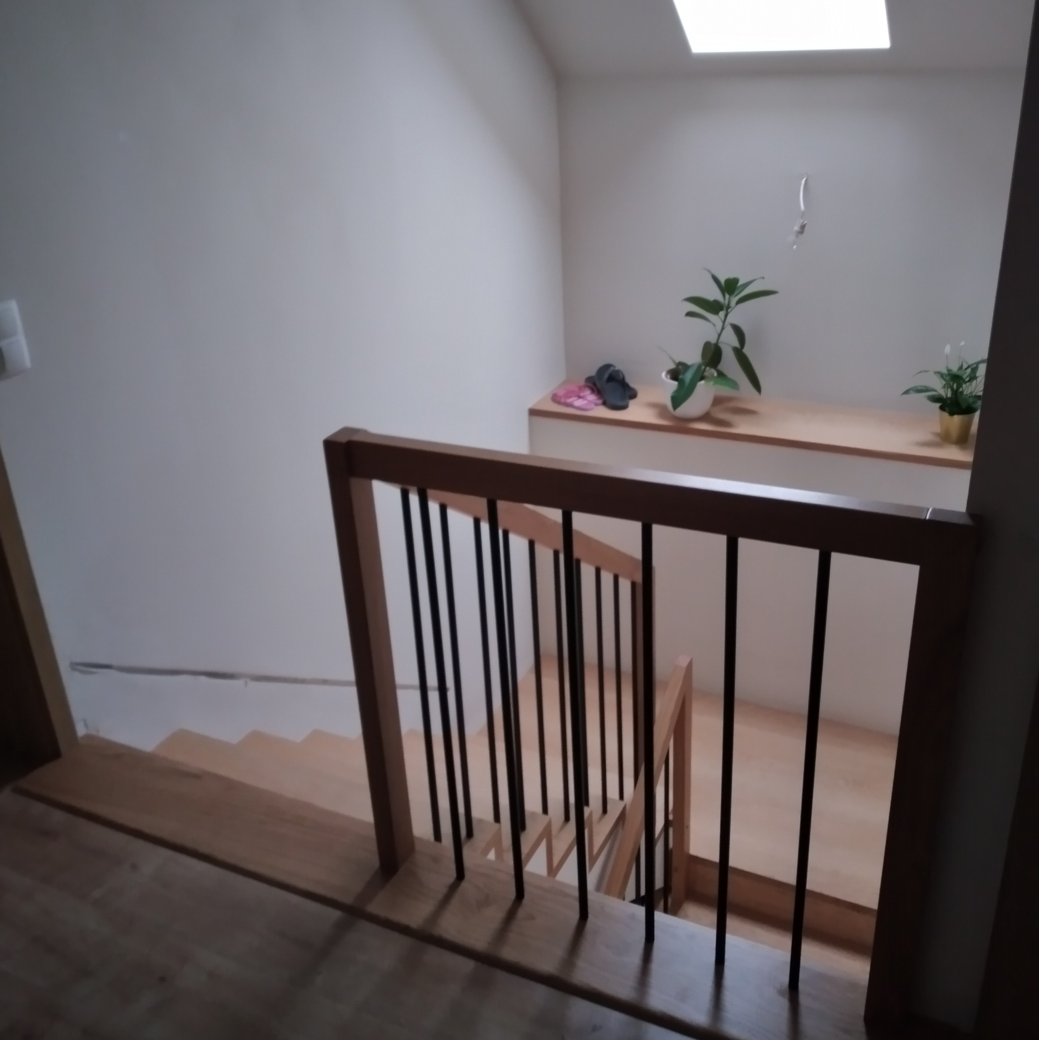 Widok z góry na jasne drewniane schody z czarnymi, pionowymi balustradami, z półką na górze z roślinami i kapciami, oświetlone przez świetlik.