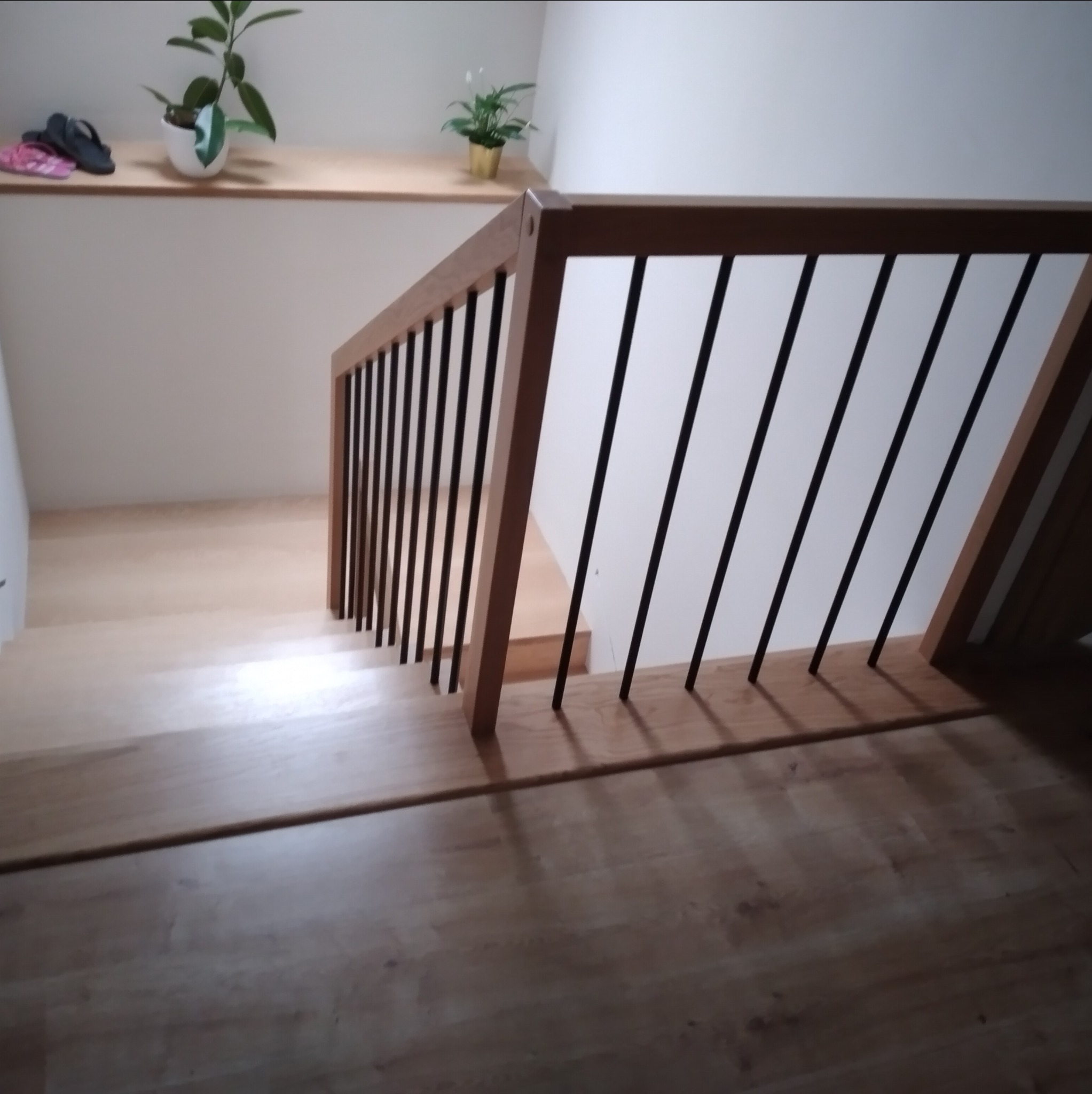 Wnętrze domu z jasnymi, drewnianymi schodami i balustradą z drewnianą ramą i czarnymi, pionowymi szczeblami. Na półce nad schodami widoczne rośliny doniczkowe i klapki.