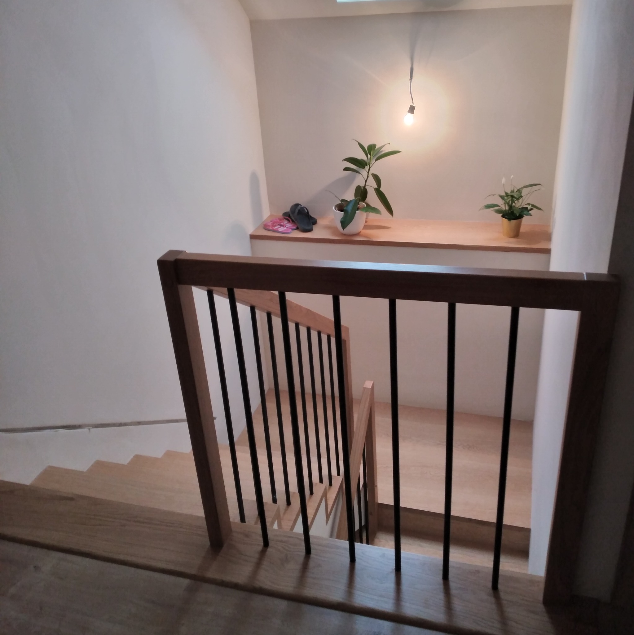 Klatka schodowa z drewnianymi schodami i balustradą z drewnianą ramą i czarnymi, pionowymi prętami. Na górze półka z roślinami i lampką.