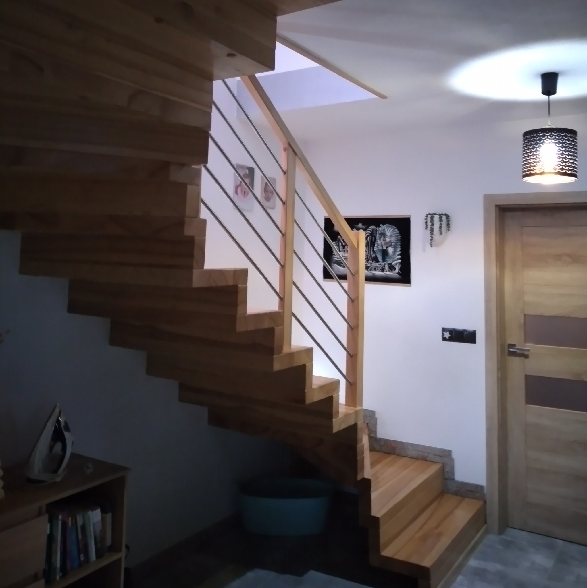 Drewniane schody z metalową balustradą w nowoczesnym wnętrzu, oświetlone lampą z ażurowym abażurem, widoczne zdjęcia na ścianie i dekoracje.