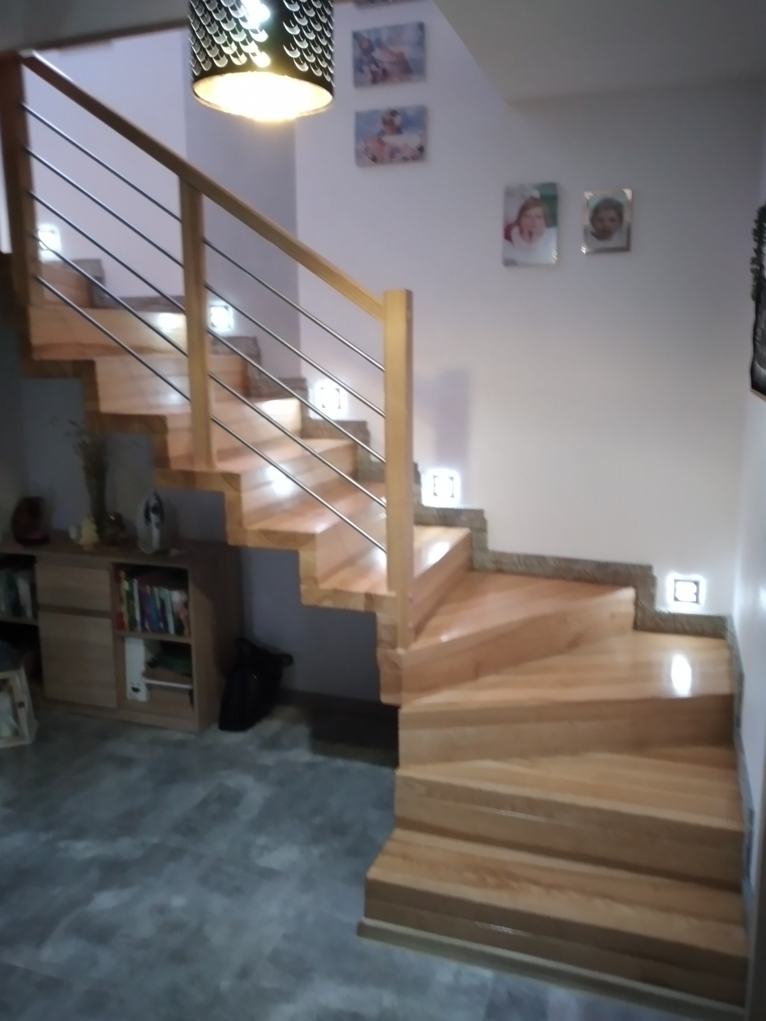 Jasne, drewniane schody z metalową balustradą i podświetleniem LED w ścianie, prowadzące na górę, z widoczną szafką i dekoracjami w tle.