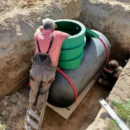 Montaż przydomowej oczyszczalni ścieków: Dwóch mężczyzn instaluje zbiornik w wykopie, jeden na drabinie sprawdza połączenie, drugi wewnątrz wykopu mocuje elementy.