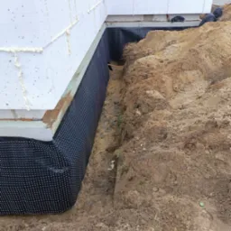 Ocieplony fundament budynku z widoczną czarną folią izolacyjną, wykopany rów zasypywany ziemią, ściany z białych bloczków.