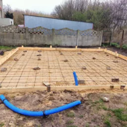 Przygotowanie terenu pod wylewkę fundamentów: drewniany szalunek, siatka zbrojeniowa podparta cegłami, niebieskie rury kanalizacyjne na ziemi.