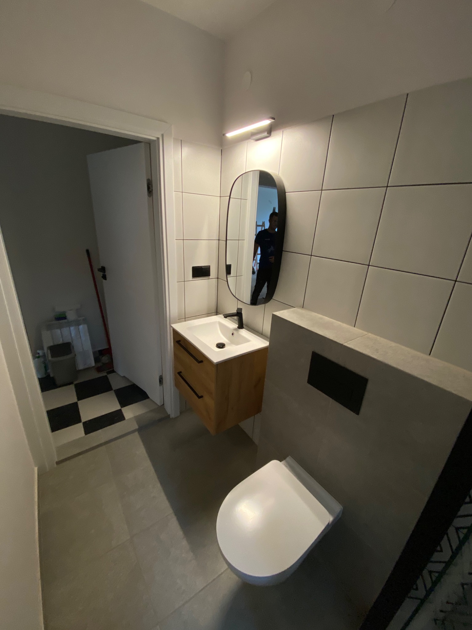 Nowoczesna łazienka z wiszącą szafką z umywalką, owalnym lustrem i podwieszaną toaletą, wykończona płytkami w odcieniach szarości.