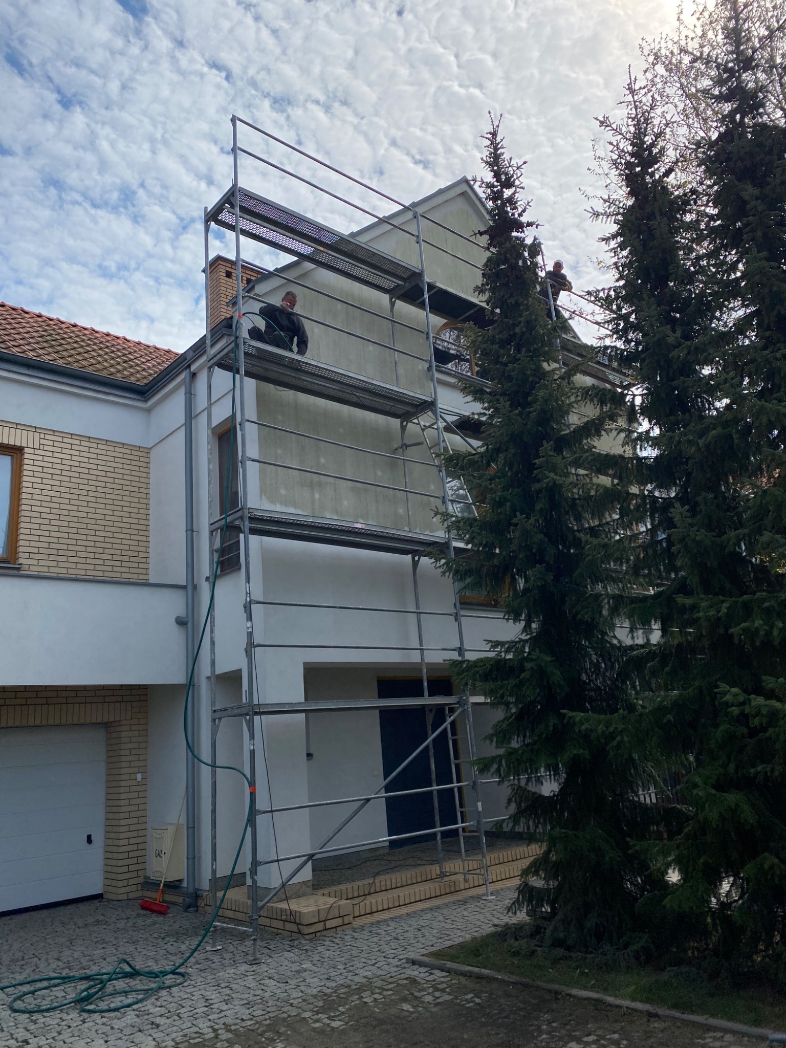 Dwóch pracowników na rusztowaniu przy dwupiętrowym domu z ceglaną elewacją, przygotowujących ścianę do malowania; widoczne drzewa iglaste i błękitne niebo.