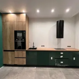 Nowoczesna kuchnia z zielonymi szafkami na wysoki połysk, drewnianymi szafkami wiszącymi i czarnym okapem cylindrycznym, widoczne zabezpieczenia folią na frontach szuflad.