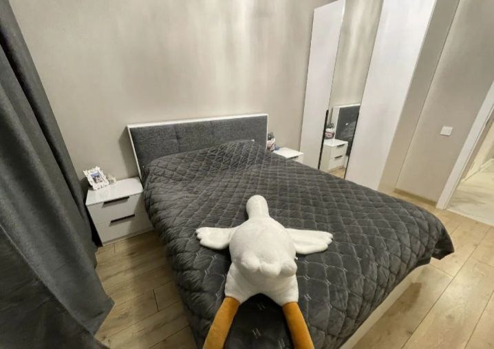 Sypialnia w odcieniach szarości z pikowaną narzutą i pluszową kaczką leżącą na łóżku, widoczne białe szafki nocne i wysokie lustrzane drzwi szafy.