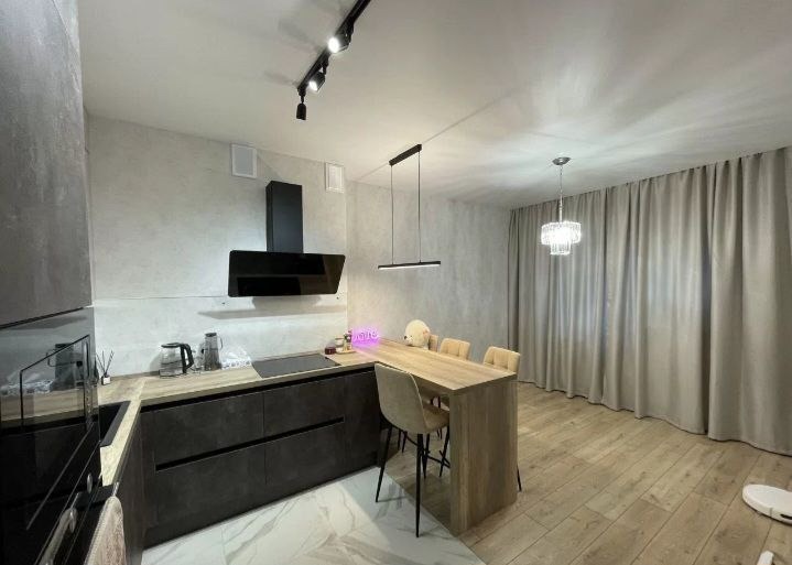 Nowoczesna kuchnia z szarymi ścianami o fakturze betonu, ciemnymi szafkami, drewnianym blatem i wyspą z krzesłami barowymi, oświetlona lampami sufitowymi i wiszącą lampą nad wyspą, z widocznym...