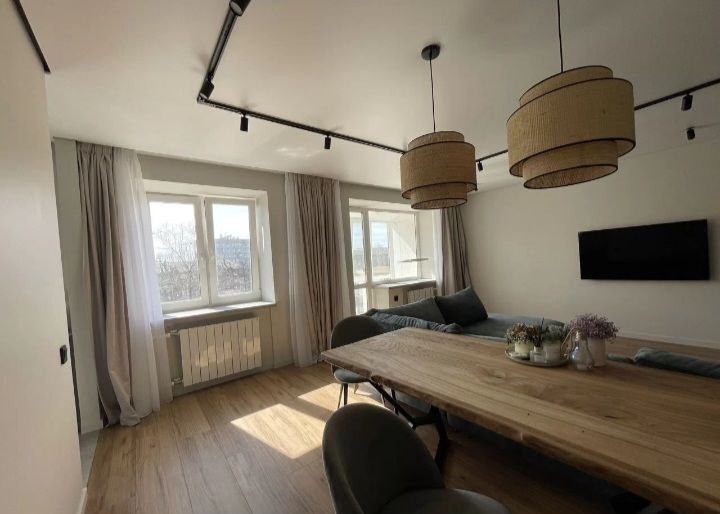 Jasne wnętrze pokoju dziennego z drewnianą podłogą, dużym stołem z surowego drewna, szarą sofą, dwoma oknami z beżowymi zasłonami i lampami z abażurami z naturalnego materiału, oświetleniem...