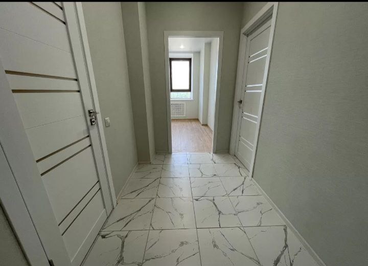 Korytarz po remoncie z białymi drzwiami z poziomymi frezami i podłogą wyłożoną białymi płytkami imitującymi marmur z szarymi żyłami, widok w głąb mieszkania przez otwarte drzwi.