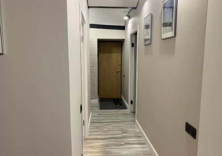 Wykończony korytarz w mieszkaniu z panelami podłogowymi imitującymi drewno, szarą ścianą z dwoma obrazami w czarnych ramkach i drewnianymi drzwiami wejściowymi.