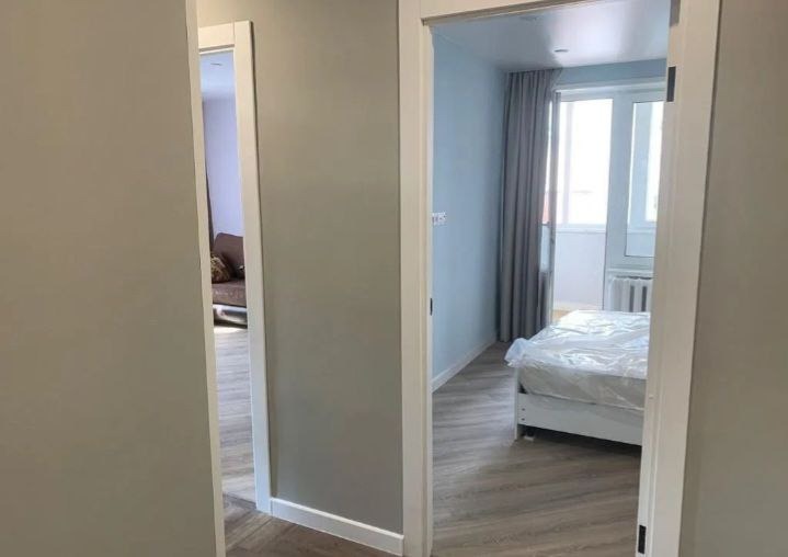 Widok na dwa pokoje po remoncie z nowymi, białymi ościeżnicami drzwiowymi i szarymi ścianami. W pokoju po prawej widoczne łóżko z pościelą zabezpieczoną folią, szare zasłony i grzejnik. W pokoju po...