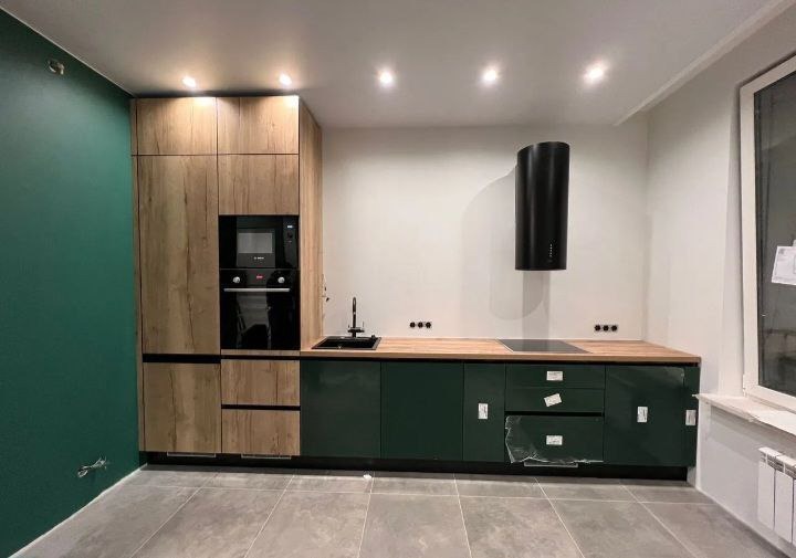 Nowoczesna kuchnia z zielonymi szafkami na wysoki połysk, drewnianymi szafkami wiszącymi i czarnym okapem cylindrycznym, widoczne zabezpieczenia folią na frontach szuflad.