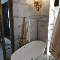 JMB-BUD JAKUB OSUCH - Nowoczesna łazienka z wolnostojącą białą wanną, złotą armaturą i dekoracyjną lampą wiszącą w kolorze złota na tle marmurowych ścian z podświetleniem.