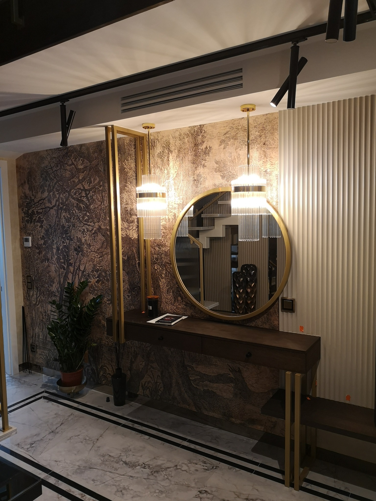 Nowoczesny przedpokój z tapetą w leśny motyw, okrągłym lustrem w złotej ramie, konsolą z szufladą, dwiema wiszącymi lampami z transparentnymi elementami i marmurową podłogą z czarnymi paskami.