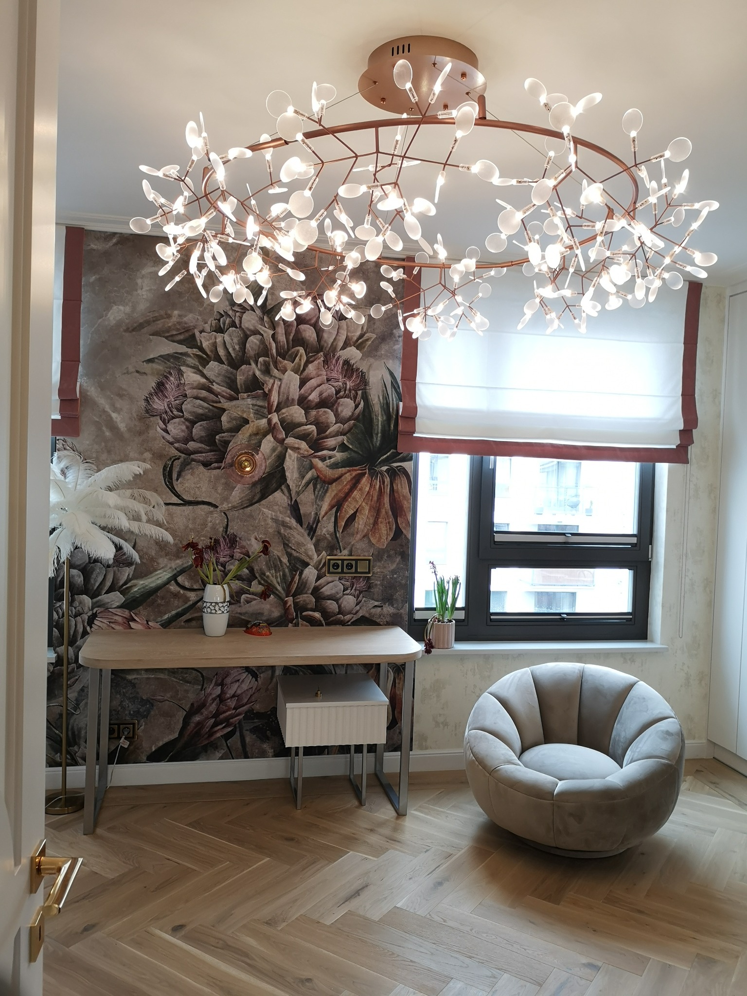 Pokój z nowoczesnym oświetleniem sufitowym w kształcie rozgałęzionych gałęzi z białymi listkami, biurkiem z dekoracyjnym wazonem na tle ściany z tapetą w kwiaty, oraz szarym, okrągłym fotelem obok...