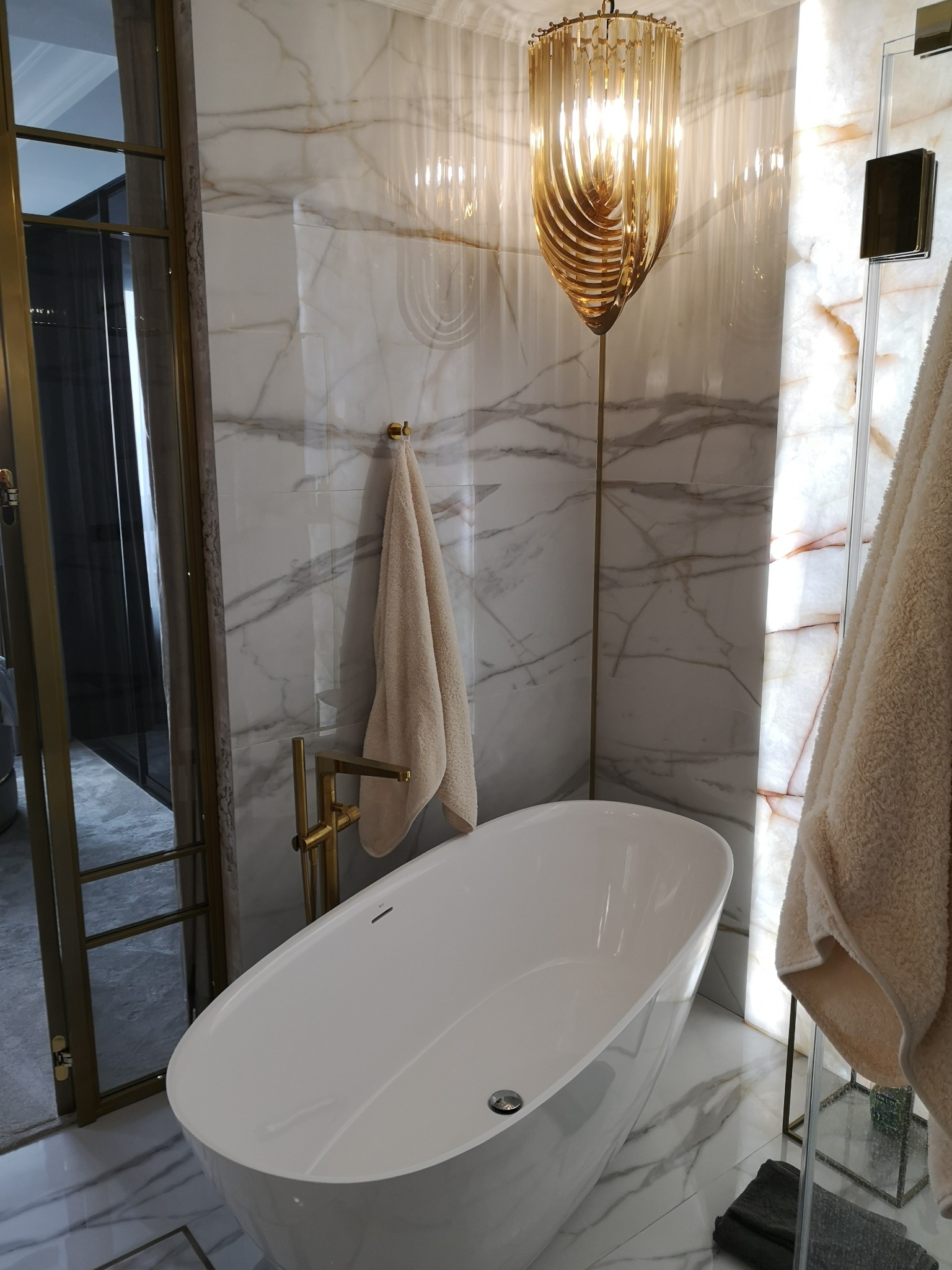Nowoczesna łazienka z wolnostojącą białą wanną, złotą armaturą i dekoracyjną lampą wiszącą w kolorze złota na tle marmurowych ścian z podświetleniem.