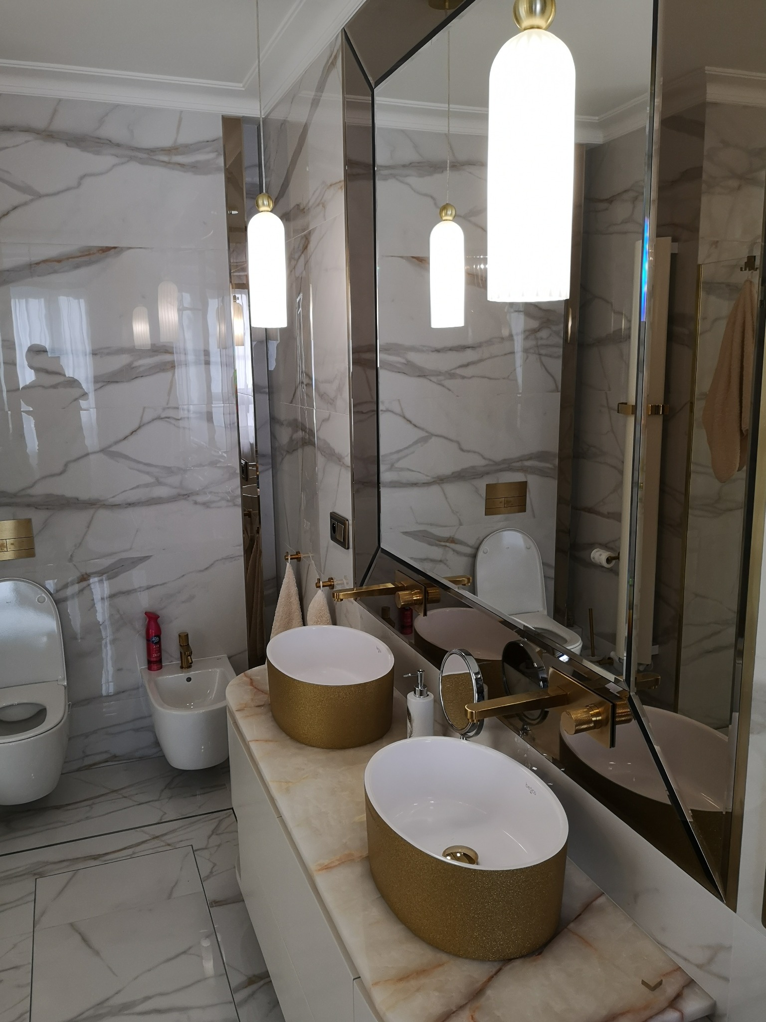Luksusowa łazienka z marmurowymi płytkami, złotymi akcentami i owalnymi umywalkami nablatowymi, oświetlona wiszącymi lampami w kształcie tub. Widoczne toaleta, bidet i duże lustro z geometrycznymi...