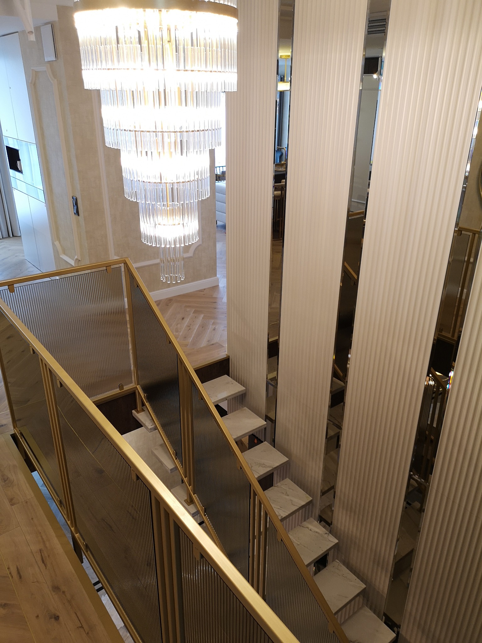 Nowoczesne schody z marmurowymi stopniami i złotą balustradą z ryflowanego szkła, oświetlone wielopoziomowym kryształowym żyrandolem, z widocznymi pionowymi panelami ściennymi z lustrzanymi...