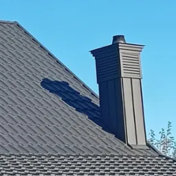 Szary dach z metalowej dach&oacute;wki z ciemnym kominem z widocznymi elementami wentylacyjnymi na tle błękitnego nieba.