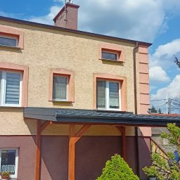 Marcin Małkowski - Dwukondygnacyjny dom z beżową elewacją i różowymi obramowaniami okien, z ciemnoszarą zadaszoną werandą wspartą na drewnianych słupach, widoczny fragment dachu i komin, na tle błękitnego nieba...