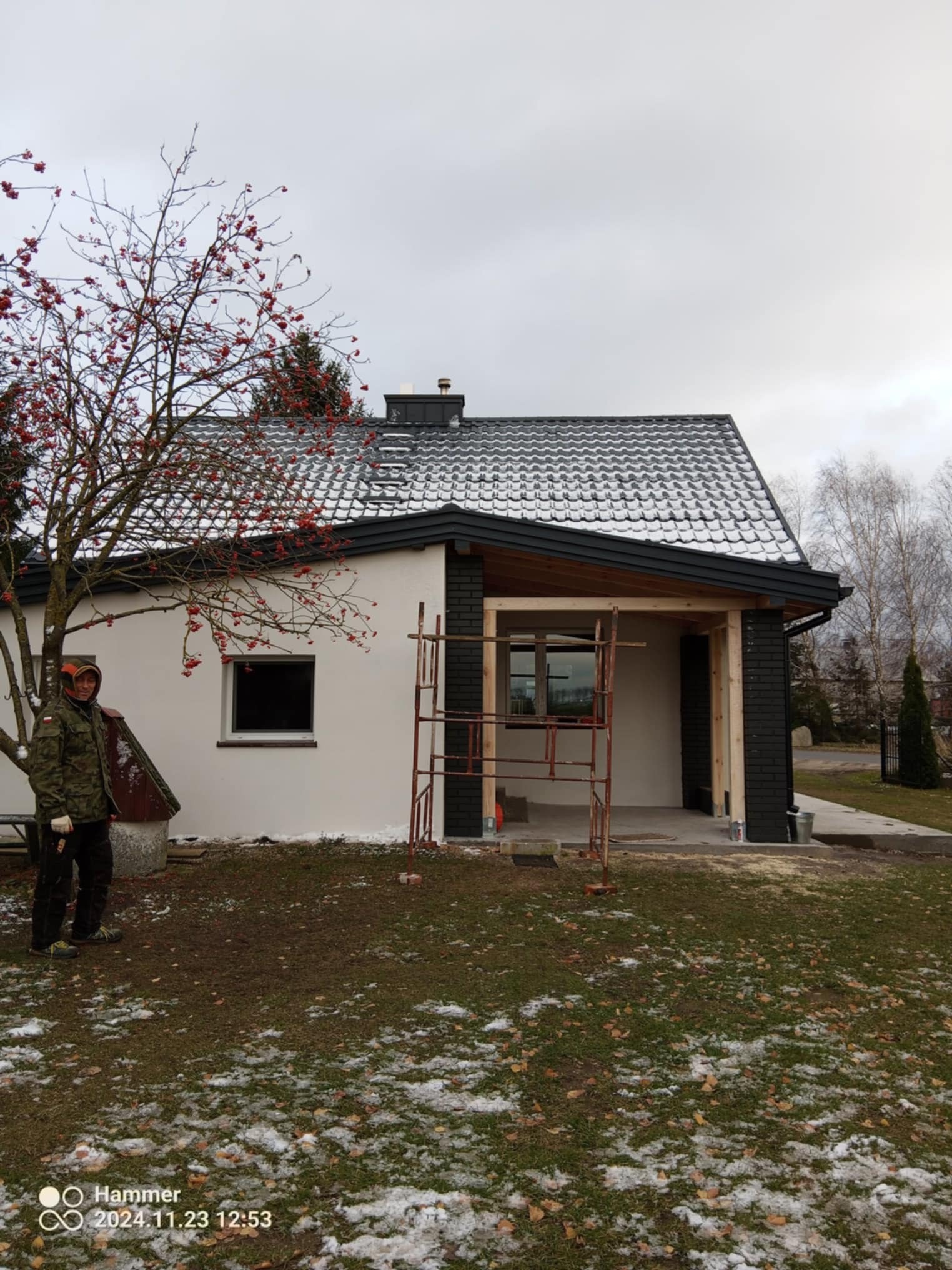Dom jednorodzinny w Lidzbarku w trakcie remontu, widoczne rusztowanie, ocieplona elewacja w kolorze białym i czarnym, nowy dach pokryty śniegiem, pracownik w ubraniu roboczym stoi obok drzewa...