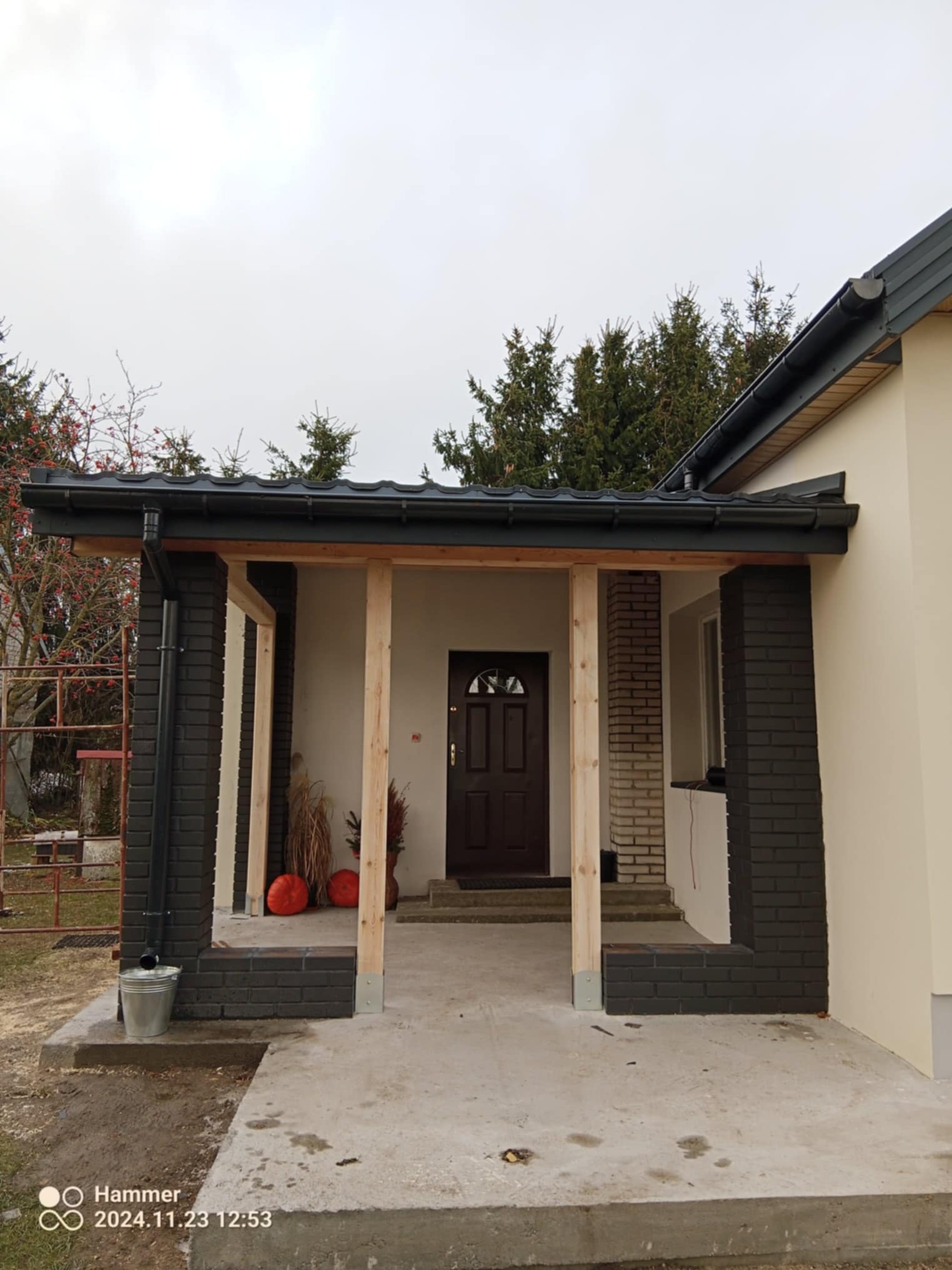 Zadaszone wejście do domu w trakcie budowy, widoczne surowe drewniane podpory, częściowo wykończone ceglane filary pomalowane na ciemny kolor, metalowa rynna i wiadro budowlane przy podstawie filaru.