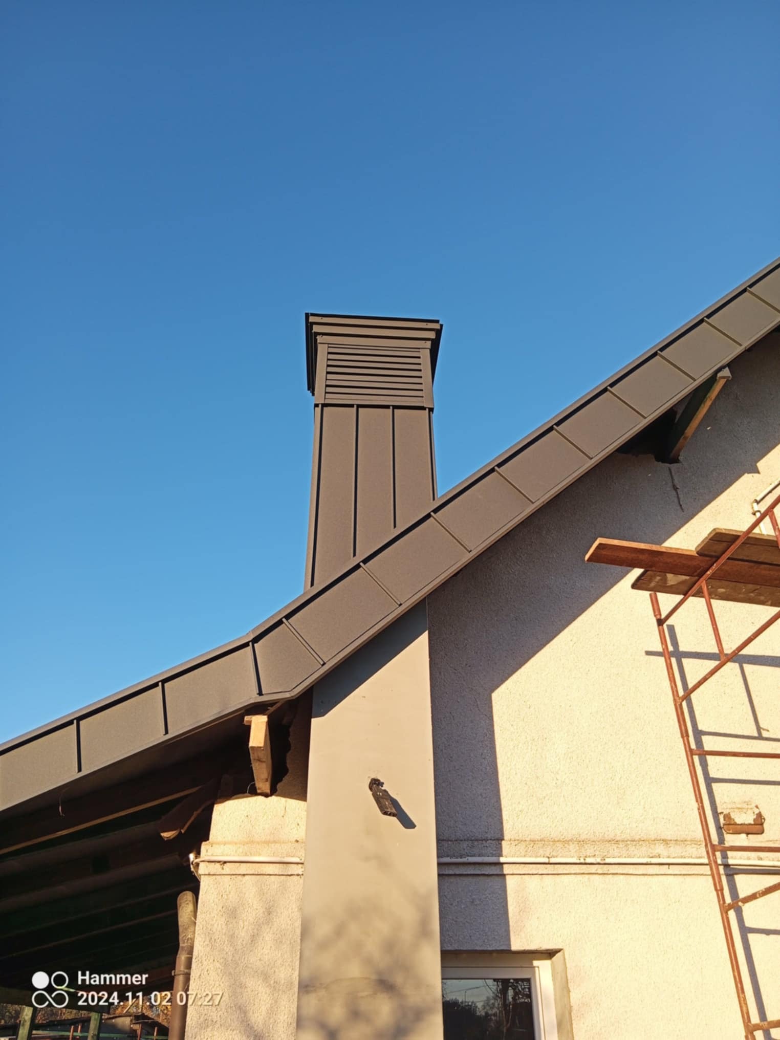 Nowoczesny, grafitowy dach z blachy trapezowej i komin w tym samym kolorze, widoczne rusztowanie przy ścianie budynku, błękitne niebo w tle.