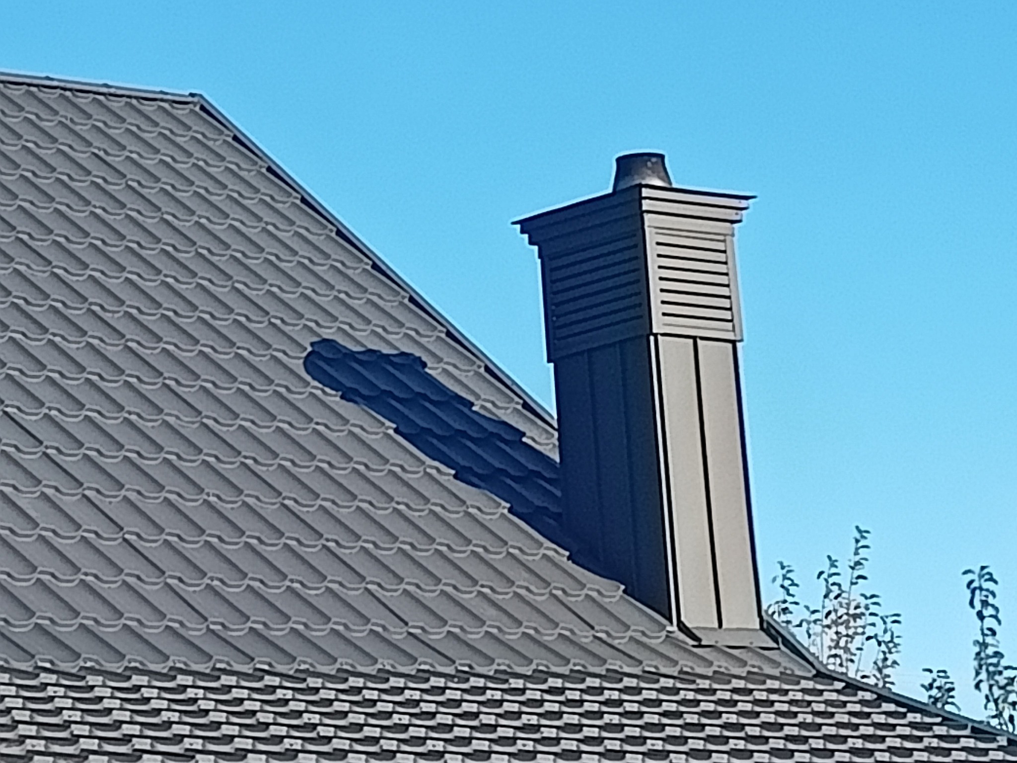 Szary dach z metalowej dachówki z ciemnym kominem z widocznymi elementami wentylacyjnymi na tle błękitnego nieba.