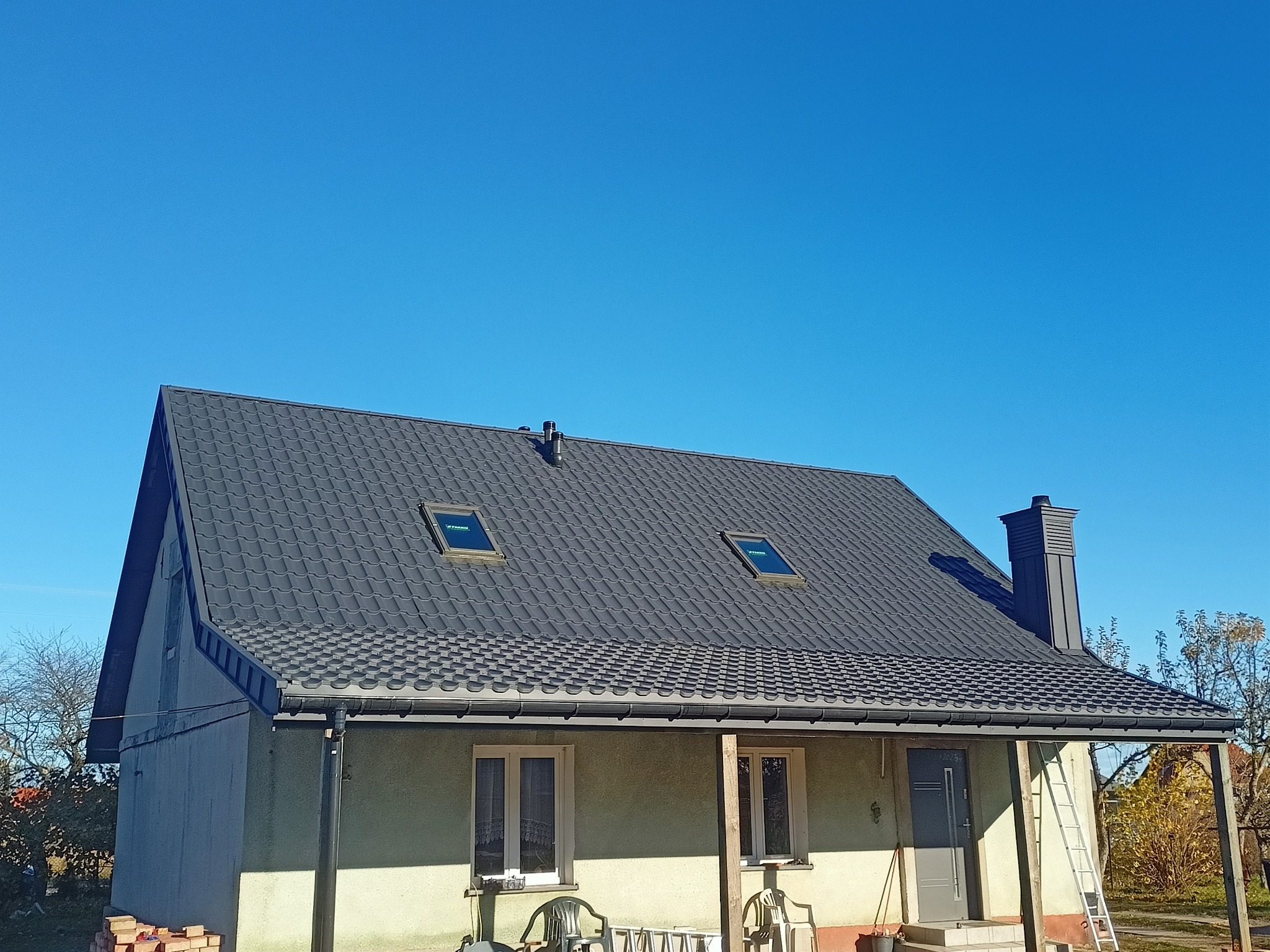 Dom jednorodzinny z nowym, ciemnoszarym dachem z blachodachówki, dwoma oknami dachowymi i orynnowaniem, na tle błękitnego nieba. Na pierwszym planie fragment zadaszonego tarasu i drabina oparta...