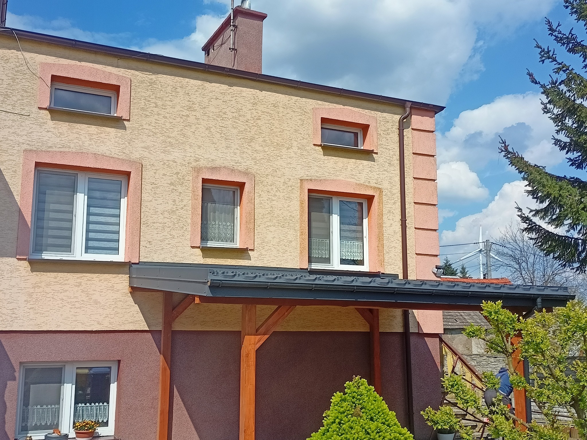 Dwukondygnacyjny dom z beżową elewacją i różowymi obramowaniami okien, z ciemnoszarą zadaszoną werandą wspartą na drewnianych słupach, widoczny fragment dachu i komin, na tle błękitnego nieba...