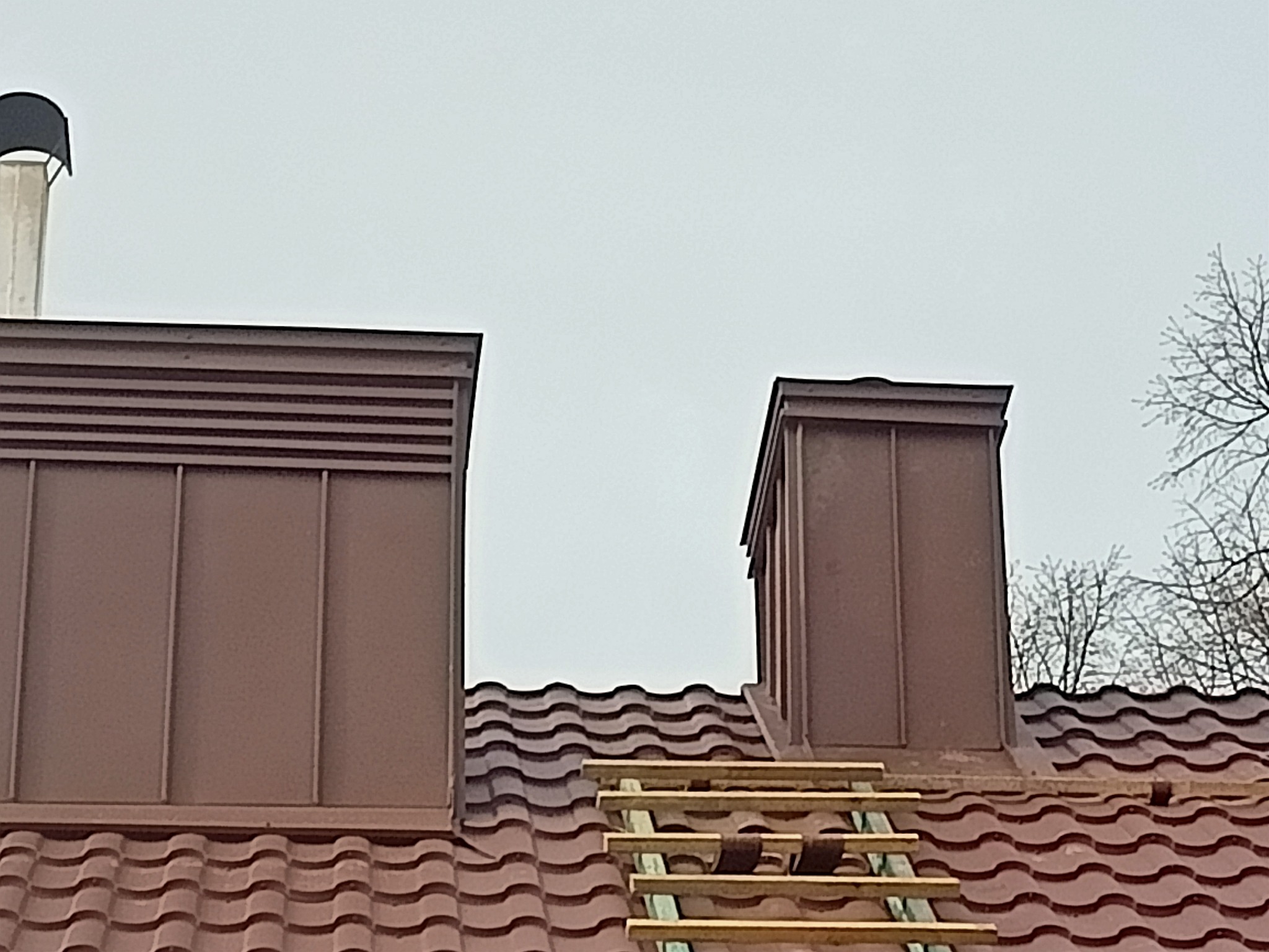 Widok z dołu na dach z brązowej dachówki ceramicznej, dwa kominy wentylacyjne w kolorze dachu oraz drewnianą drabinę dekarską opartą o połać dachu.