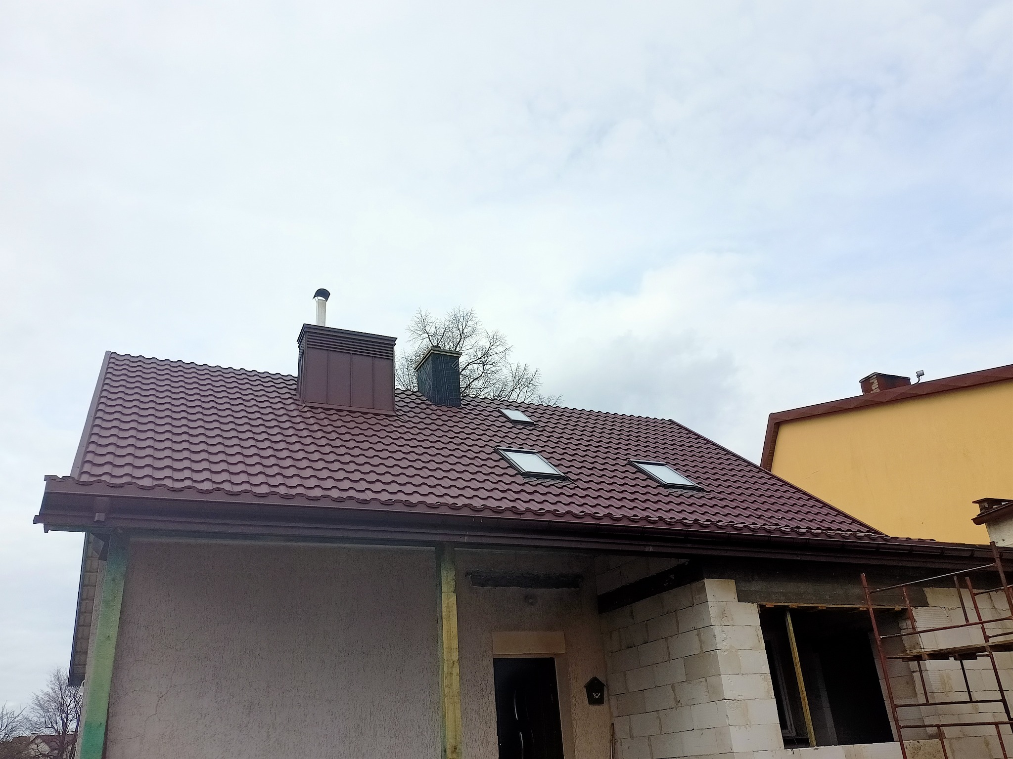 Dom w trakcie budowy z nowym, brązowym dachem z blachodachówki, trzema oknami dachowymi i brązowymi rynnami. Widoczny komin z brązową obudową i metalowym zakończeniem. Ściany budynku w stanie...