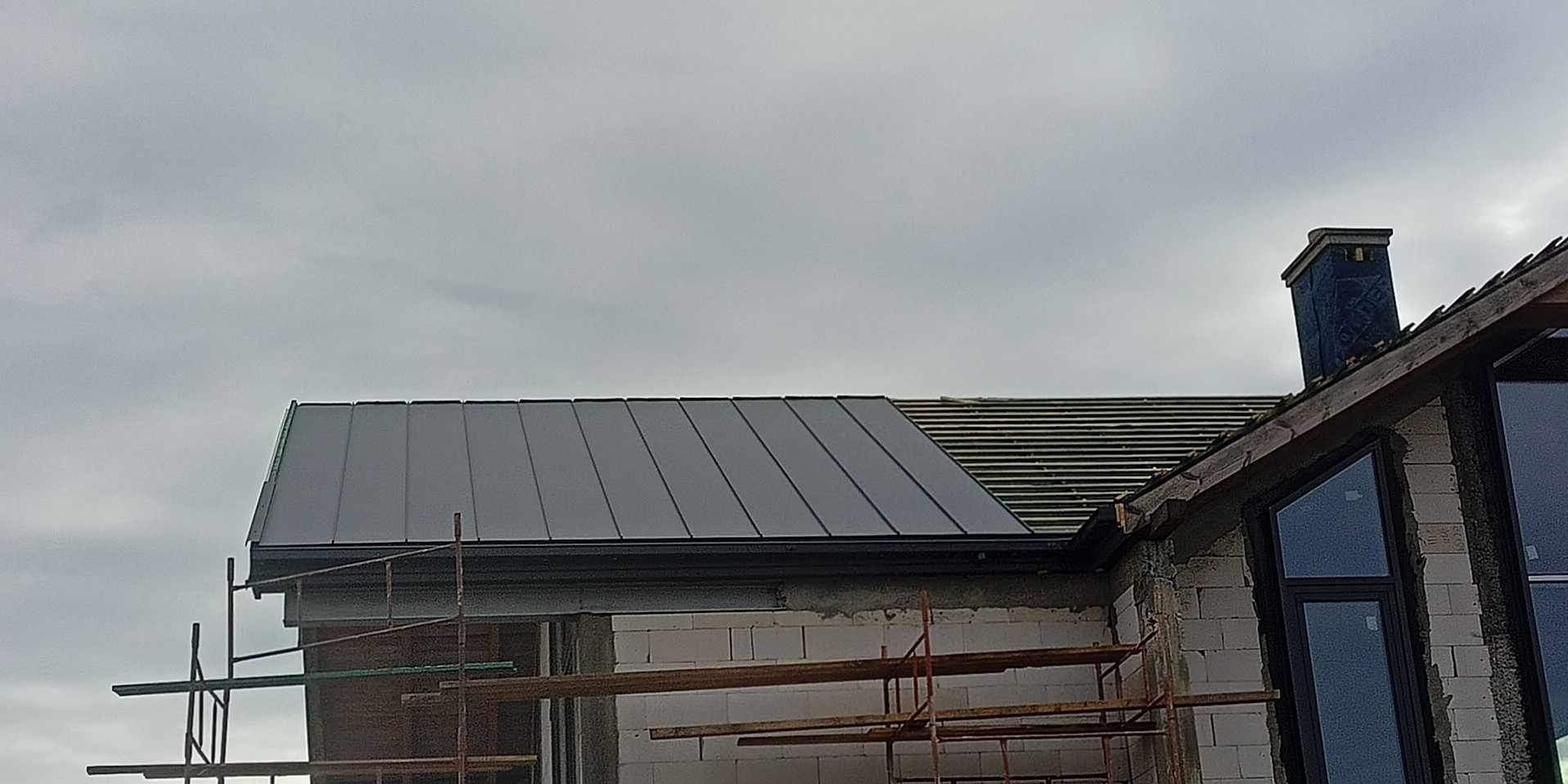 Dom w trakcie budowy z fragmentem dachu pokrytym blachą trapezową w kolorze grafitowym i częściowo ułożoną dachówką ceramiczną; widoczne rusztowania i komin w folii ochronnej.