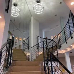 Wnętrze budynku użyteczności publicznej ze schodami wyłożonymi wykładziną, czarnymi metalowymi balustradami, lustrem na ścianie oraz nowoczesnymi, okrągłymi żyrandolami LED.