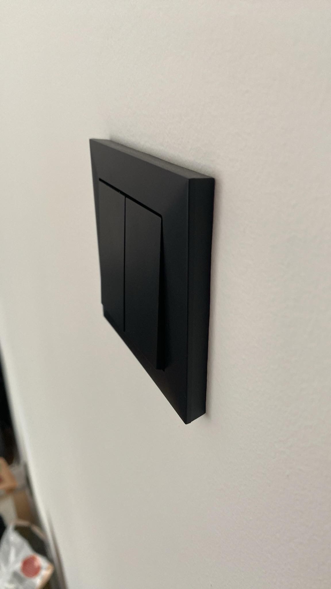 Czarny, podwójny włącznik światła z ramką, zamontowany na jasnej ścianie, widok z boku.