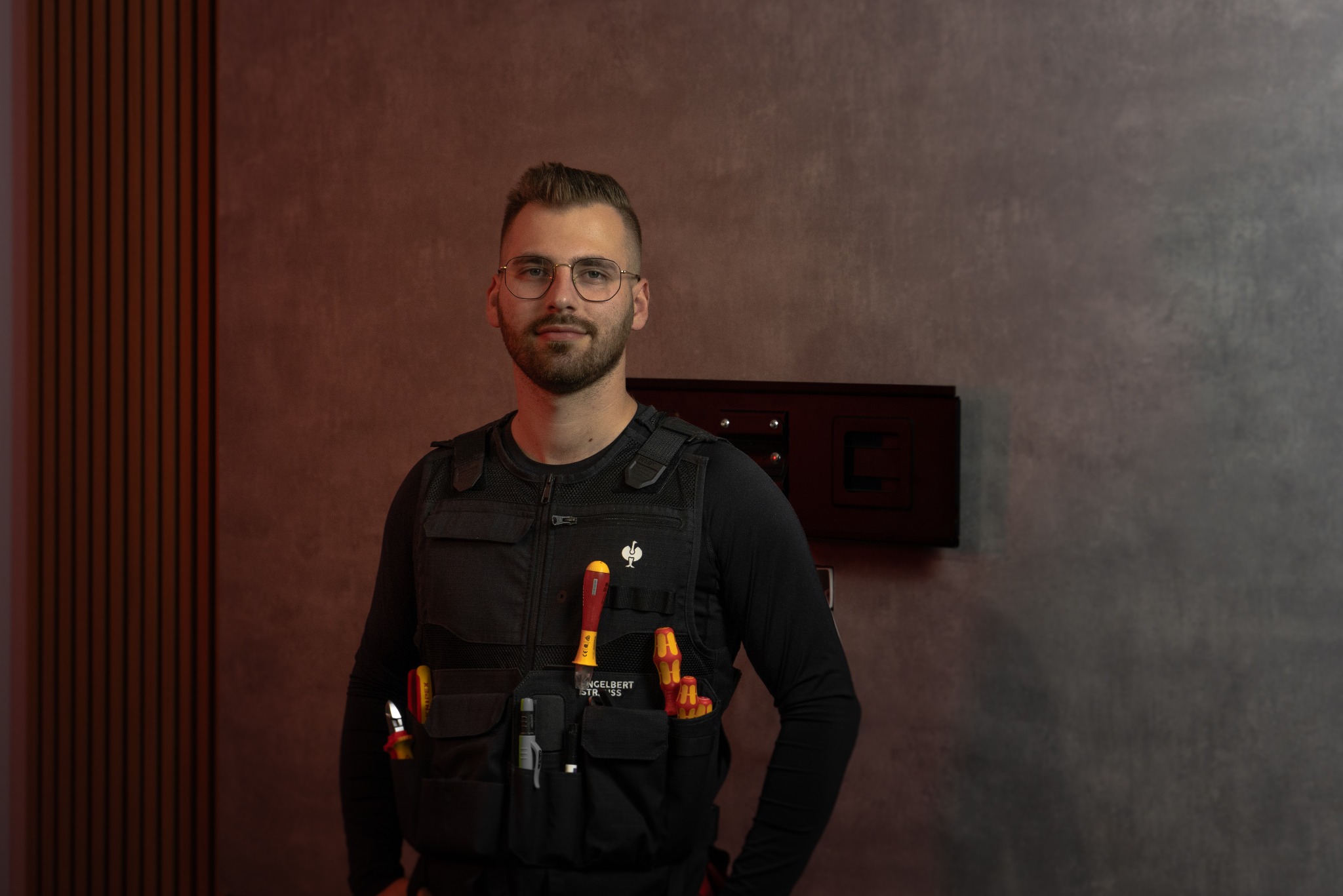 Portret elektryka w kamizelce narzędziowej Engelbert Strauss, z widoczną puszką instalacyjną na ścianie w tle
